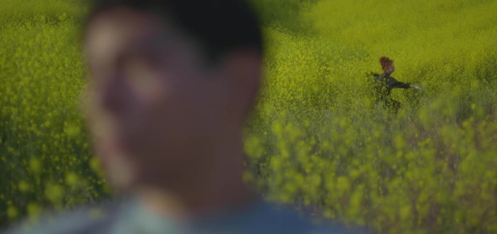 A young man standing on a grassland turning his head left and right while a woman behind him among tall grass and bushes changes her clothes at midday