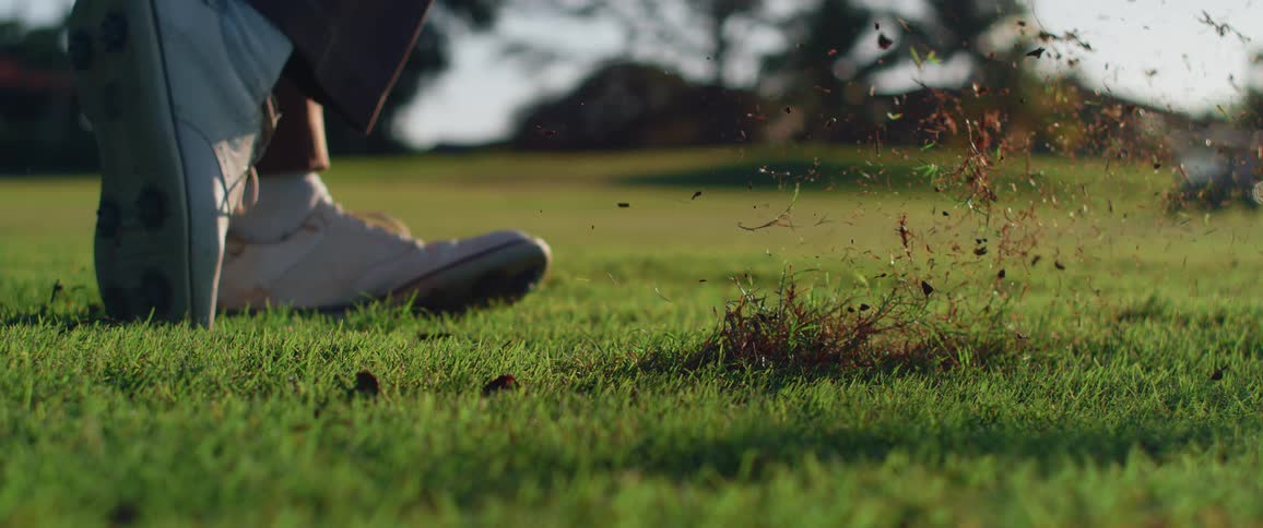 A man on a golf course wearing white shoes swings a golf club with force to hit the ball from the grass at midday