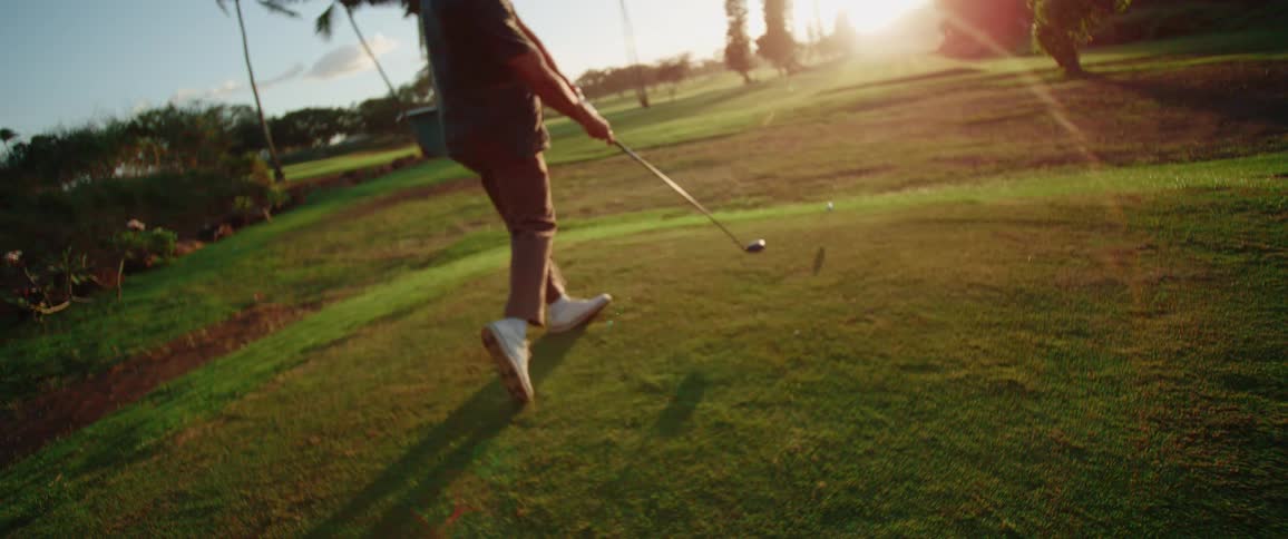 A man on a golf course wearing a hat striking a golf ball with force using a golf club at sunset