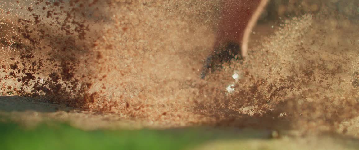 Golf club strikes the ball in the sand raising a cloud of grains