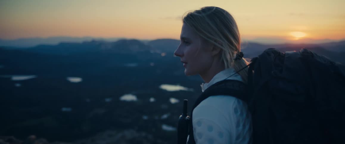 A woman with a backpack looks over mountains at sunset