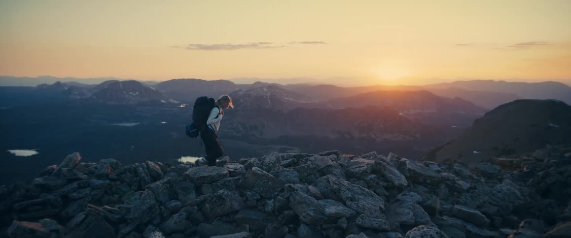White woman hiking and carrying a backpack over a mountain surrounded by a mountain range and a relatively flat area outdoors during dusk or dawn
