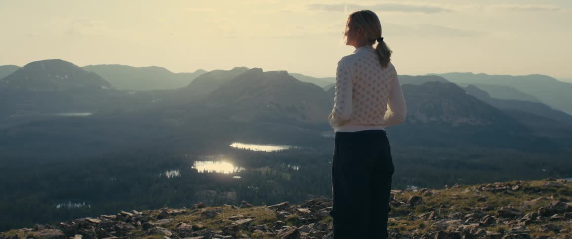 A woman stands on a mountain cliff overlooking a valley at sunset