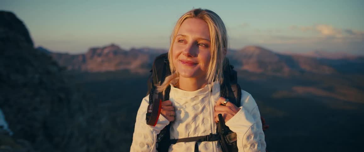 Blonde female hiker smiles while standing on a mountain and looking toward the sun