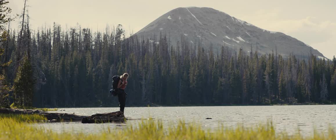 Woman carrying a backpack walking towards a body of water surounded by a forest with a mountain on the background outdoors during day time