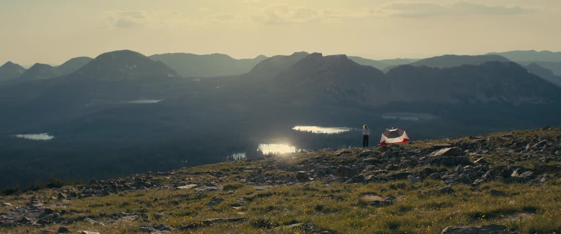 Camper stands by a tent on a mountain ridge overlooking lakes at sunset