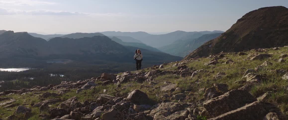 A person with a backpack walks on a slope with rocks and vegetation overlooking a landscape with mountains in the distance