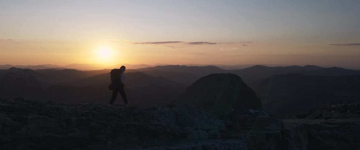 A person wearing a backpack is seen hiking along a rocky desolate path during sunrise or sunset the sun is a bright orb on the horizon silhouetting the person and a range of mountains in the background