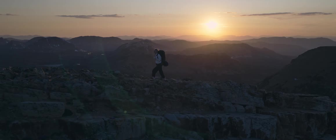 White woman carrying a backpack hiking over a rocky mountain surrounded by a mountain range during dusk or dawn