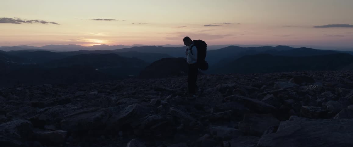 Female backpacker hikes across rock strewn terrain while the sun rises over the mountains on the horizon