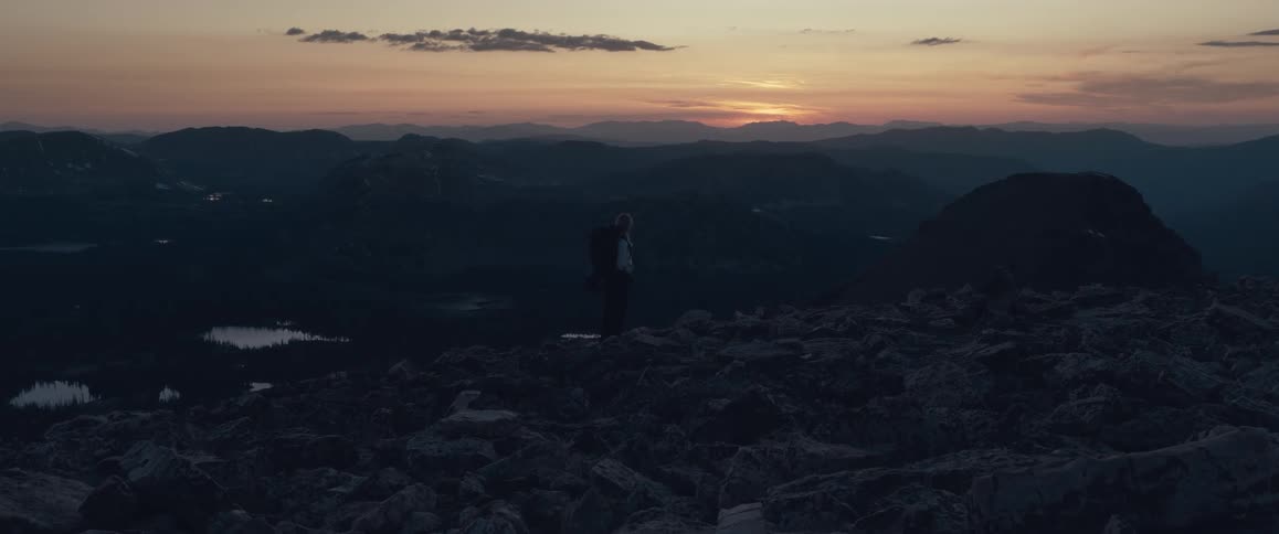 A person stands on a mountain with a sunset in the background over a mountain range