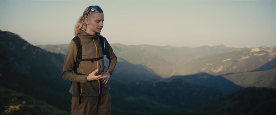 A female hiker stands on a mountainous ridge checking her phone and taking in the vast scenic landscape before she turns and continues her hike