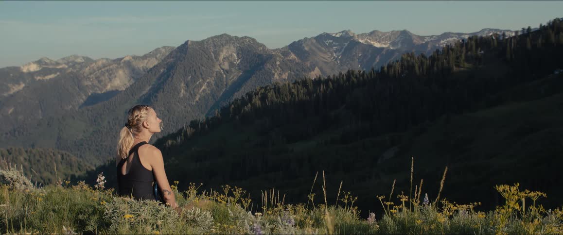 A woman with braided hair wearing a black top sits peacefully in a field of wildflowers on a mountain contemplating the view