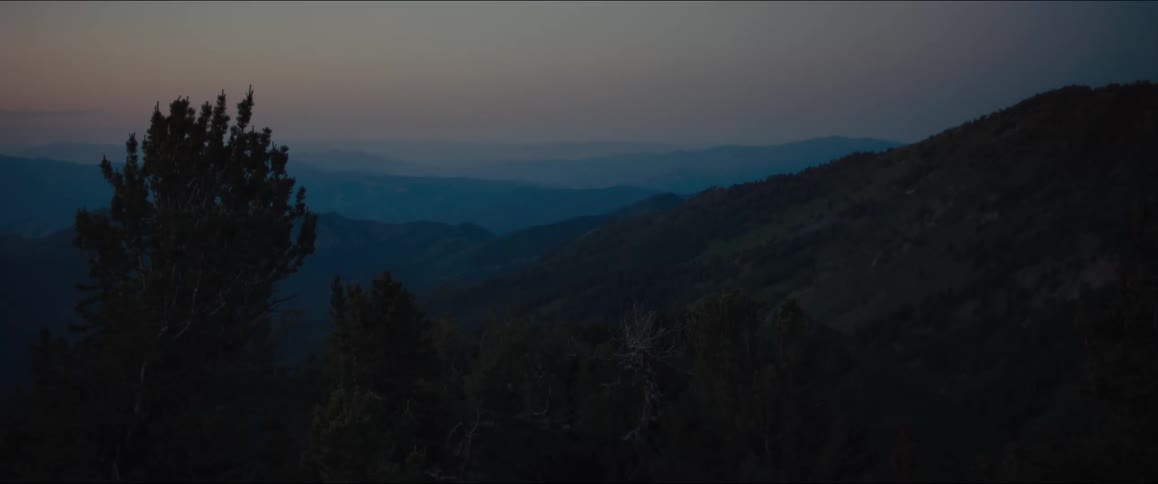 A wide shot of a hazy mountainous landscape with deep blue and purple hues with the dark silhouettes of mountain ridges and trees in the foreground