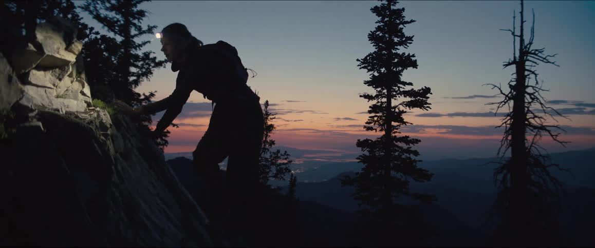 In a dark mountainous landscape a person with a headlamp and a backpack scrambles up a rocky path