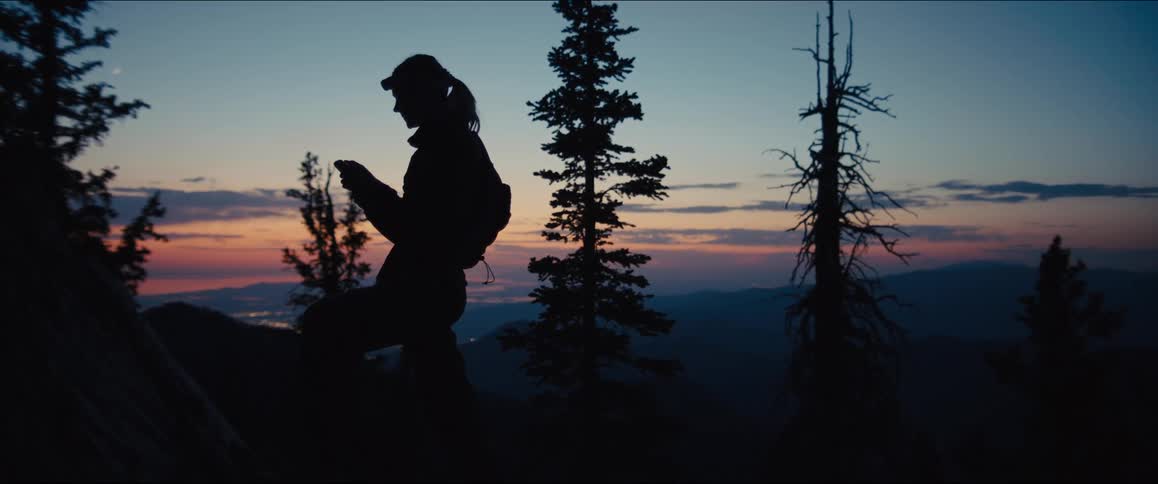 A woman stands on a mountain at dusk using a phone with a landscape in the background