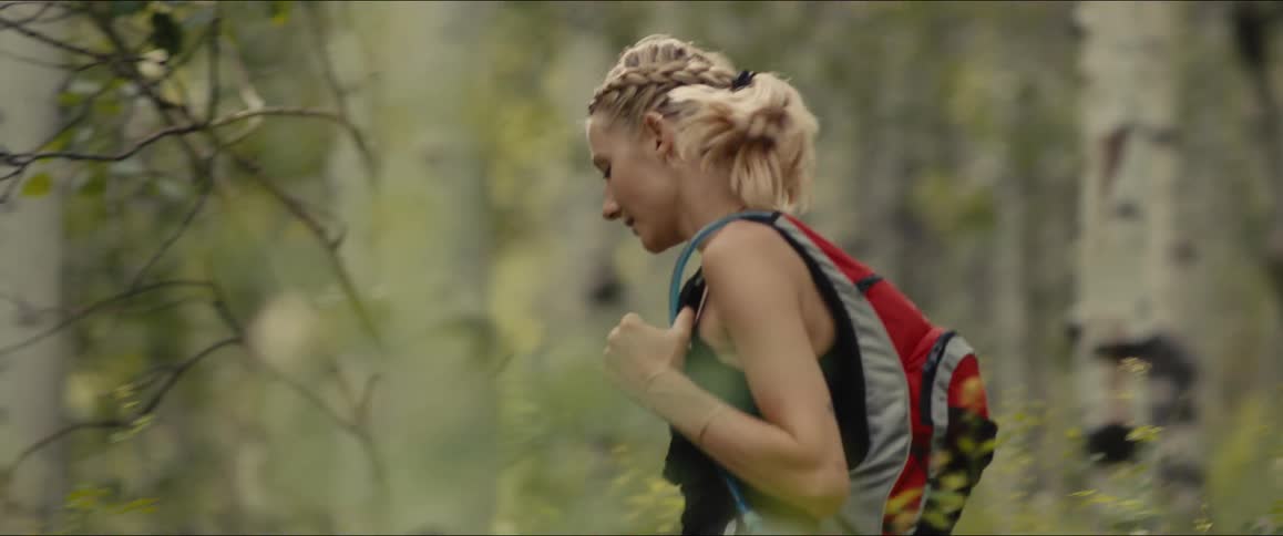 A woman with a braided ponytail and a backpack hikes through a mountainous forest filled with trees