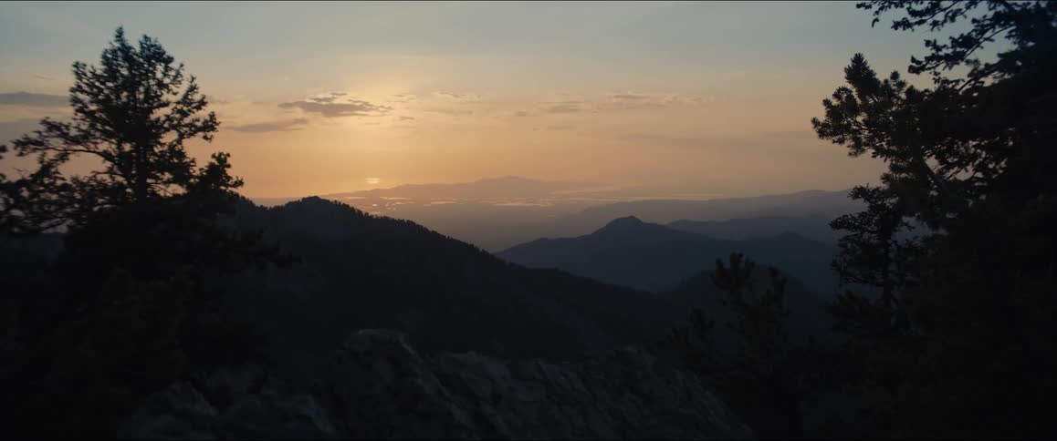 A peaceful mountainous landscape at sunset showcasing the dramatic colors of the sky and the silhouette of the mountain ridges and trees in the foreground