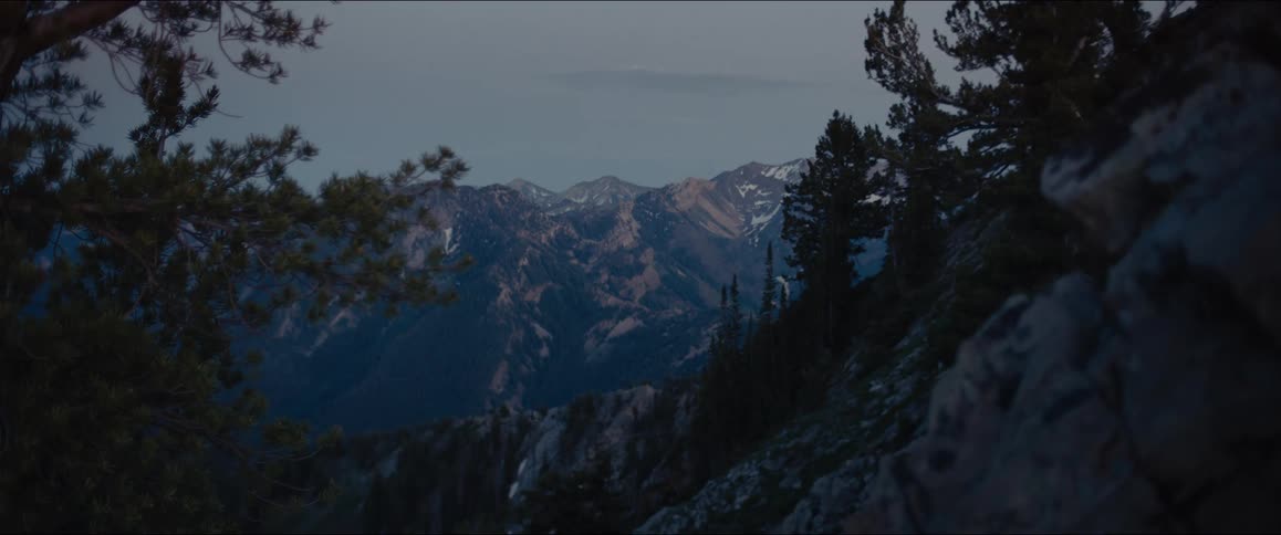 A static wide shot of a rugged mountainous landscape at dusk or dawn with snowcapped peaks in the background