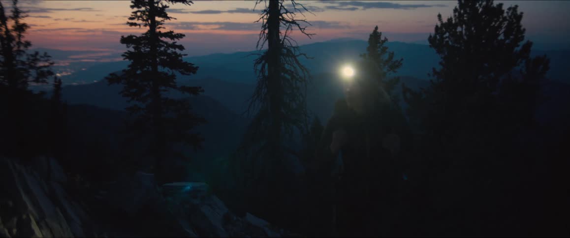 A hiker moves through a dark mountain forest with a headlamp at dawn