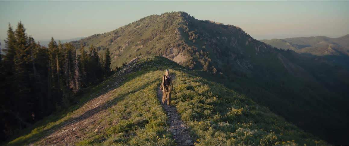 A young woman with a braided ponytail and sunglasses perched on her head hikes along a winding grassy mountain ridge overlooking a valley wearing a black tank top and brown cargo pants