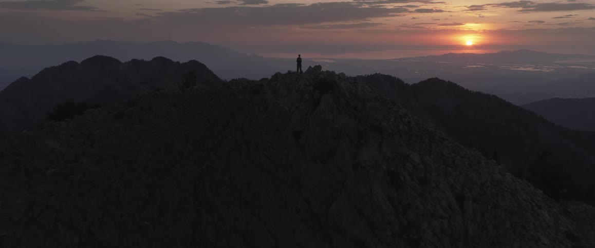 A wide shot of a person standing on a rugged mountain peak at sunset silhouetted against the sky