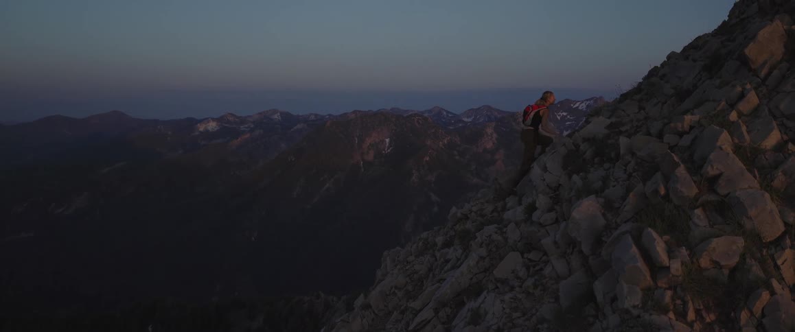 The video shows a person with a backpack carefully climbing a steep rocky mountain slope at dusk with vast mountain ranges visible in the background