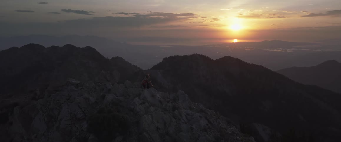 A lone person sits atop a rocky mountain peak overlooking a vast hazy mountain range as the sun sets casting a warm glow on the horizon