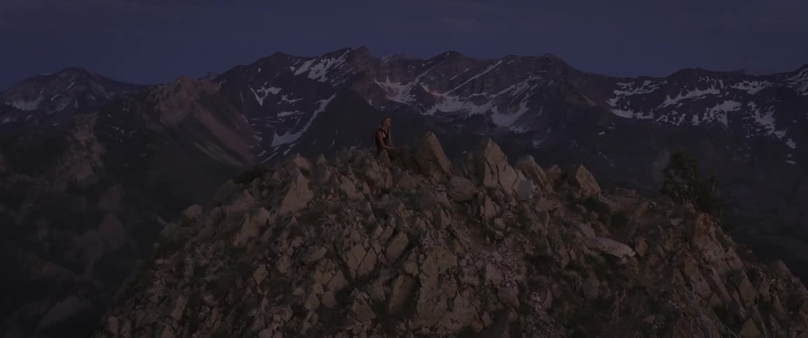 A woman sits contemplatively on a rocky mountain peak at dusk with a vast mountain range including some snowcapped peaks visible in the background