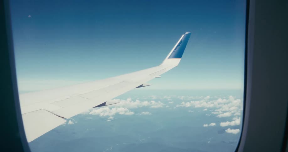A clear blue sky with scattered clouds and faint land features beneath is seen from an airplane window with the white and bluetipped wing of the plane prominently in the foreground