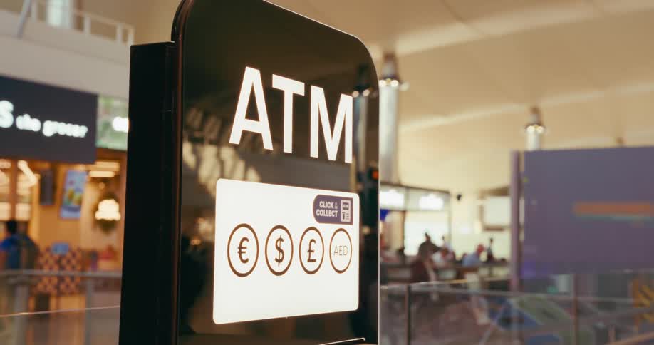 A shiny black atm sign with a white lit border stands in an open area with a white illuminated panel below it showing the symbols for euro pound dollar and aed currency