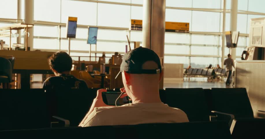 A person wearing a black baseball cap and earphones sits in an airport terminal engrossed in a video game on a handheld device while other travelers and bright windows are visible in the background