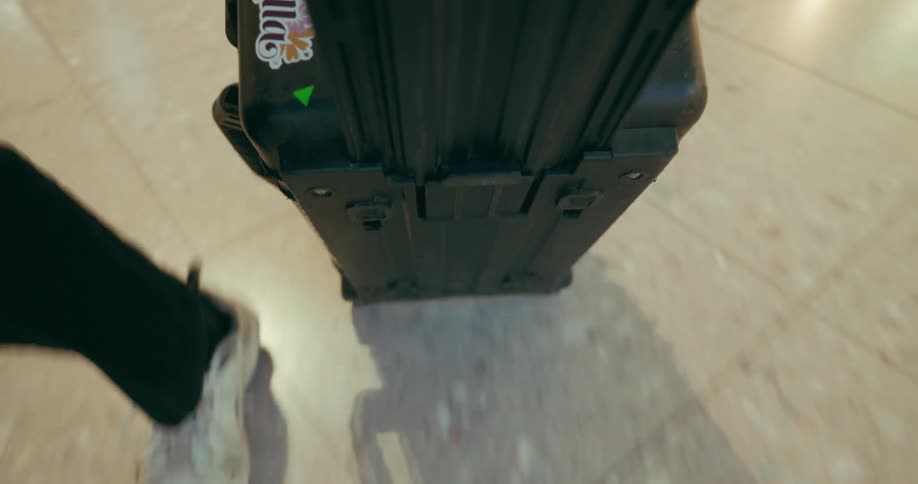 The video is shot from a low angle showing a person walking with a black hardcased rolling suitcase the camera focuses on the bottom of the suitcase which has two wheels and is being pulled on a lightcolored tile floor the persons feet wearing li