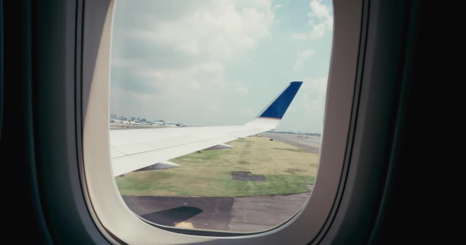 A wide view from an airplane window shows the white wing with a dark blue tail fin as the plane moves down a paved runway