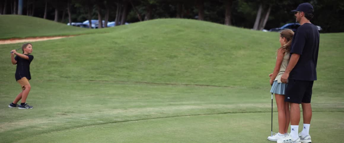 A male golf instructor with a black hat and sunglasses stands beside a young female student while another student hits a golf ball
