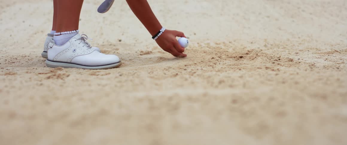 A golf ball is dropped and rolls in a sand trap a persons hand reaches in to pick it up