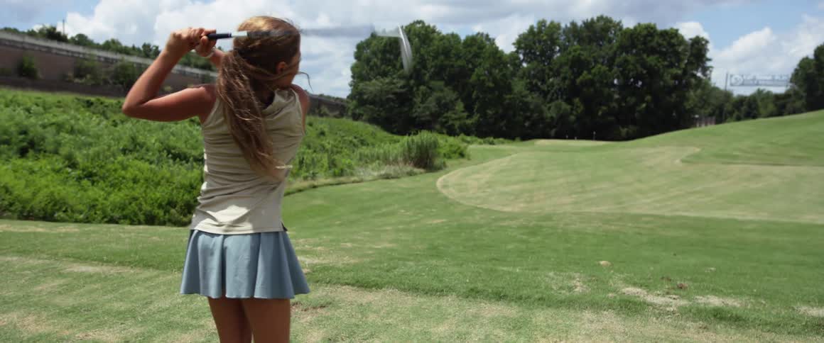 A teenage girl on a golf course swinging a golf club and hitting a golf ball