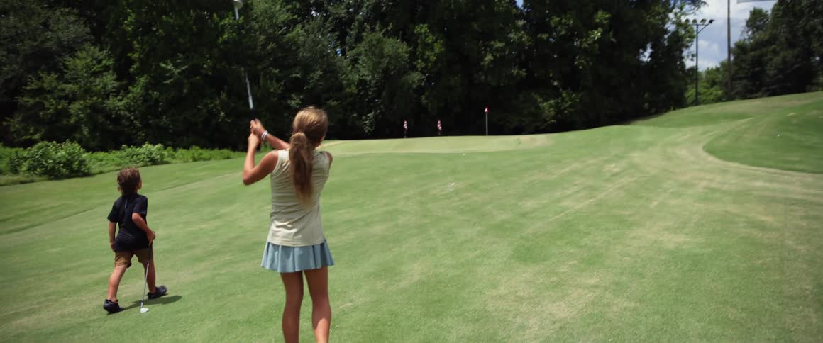 The video shows two children on a golf course a girl takes a practice swing and then hits a golf ball which can be seen flying toward the hole as her brother watches her in the background