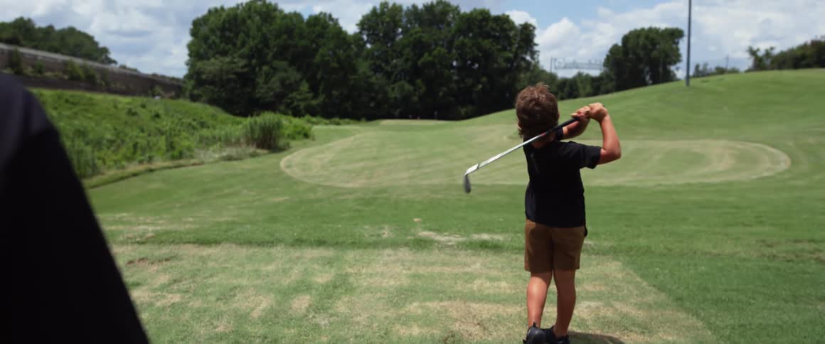 Two boys are standing on a golf course one boy is wearing a black shirt and brown shorts and is swinging a golf club another person is visible in the frame holding a golf club and standing next to him