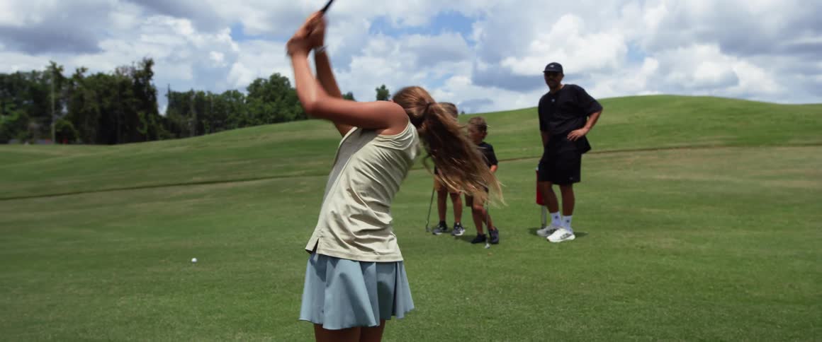 The video shows a young person with long hair pulled into a ponytail swinging a golf club they are wearing a lightcolored top and a light blue or gray skirt the video cuts to a wider shot showing the person and a group of people including two childr