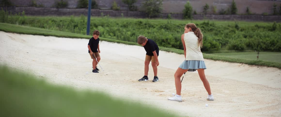 A young woman with her two kids playing golf outdoors on sand surrounded by green vegetation at midday