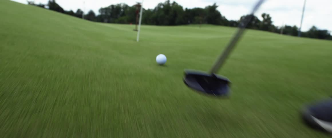 A person putts a white golf ball with a club toward a hole with a flag and the ball rolls directly into the cup