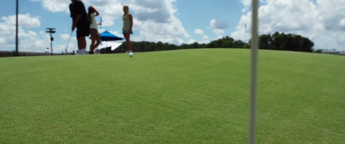 A golf ball rolls across a green golf course passing the hole with its flag while several blurred people are visible in the background under a partly cloudy sky