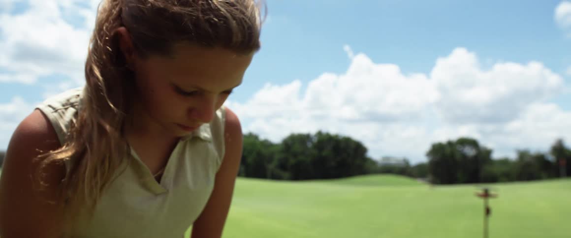The video shows a young person with long hair pulled back wearing a lightcolored polo shirt they are focused on something below the frame possibly a golf ball as a lush green golf course is visible in the background under a partly cloudy sky the per