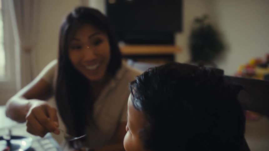 The video shows a woman possibly a mother with a warm smile using a spoon to feed a child the child is seen from the back sitting in a high chair or at a dining table the scene takes place indoors likely in a dining room
