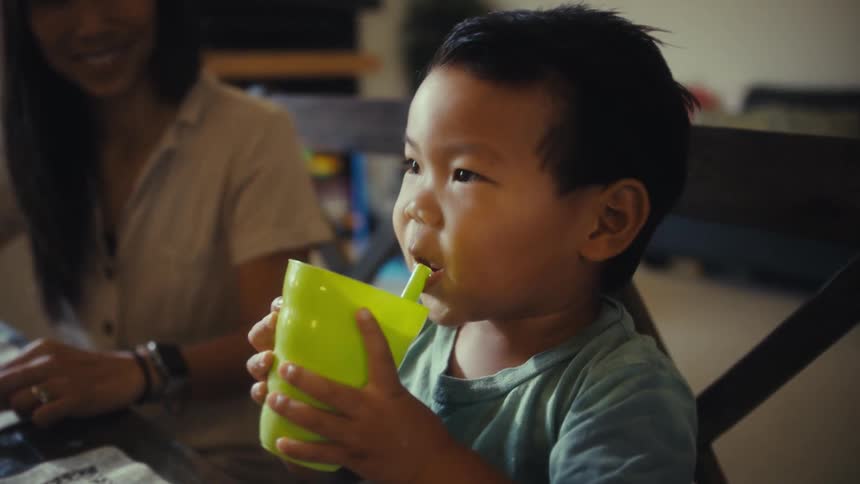 Little boy sitting at table next to adult drinking from yellow bottle with straw