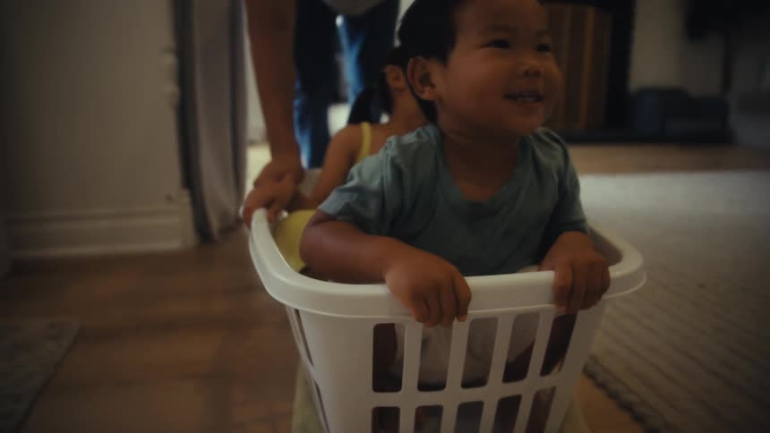 A father pushes his two young children who are seated in a white laundry basket across a wooden floor in a living room