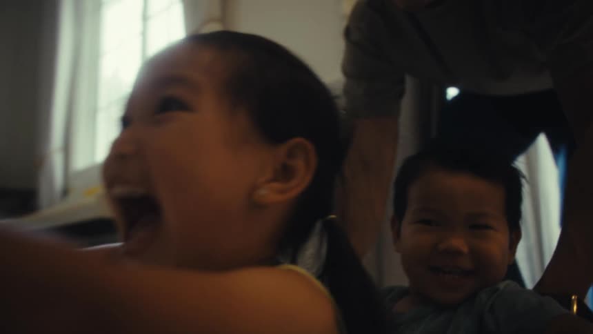 A toddler and a young girl sit inside a basket as their father playfully pushes them around the living room all enjoying a fun midday moment