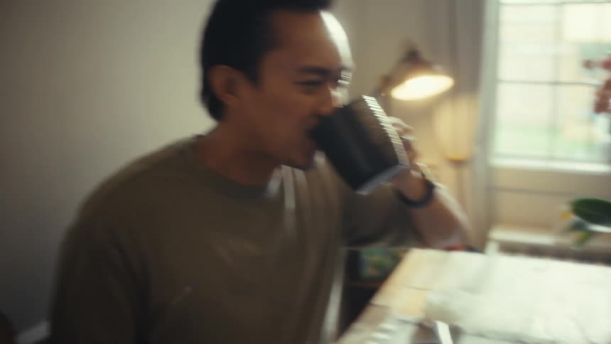 A toddler drinks from a cup and tries to hand it to his father who is also drinking from his own glass while sitting at the dining table at midday