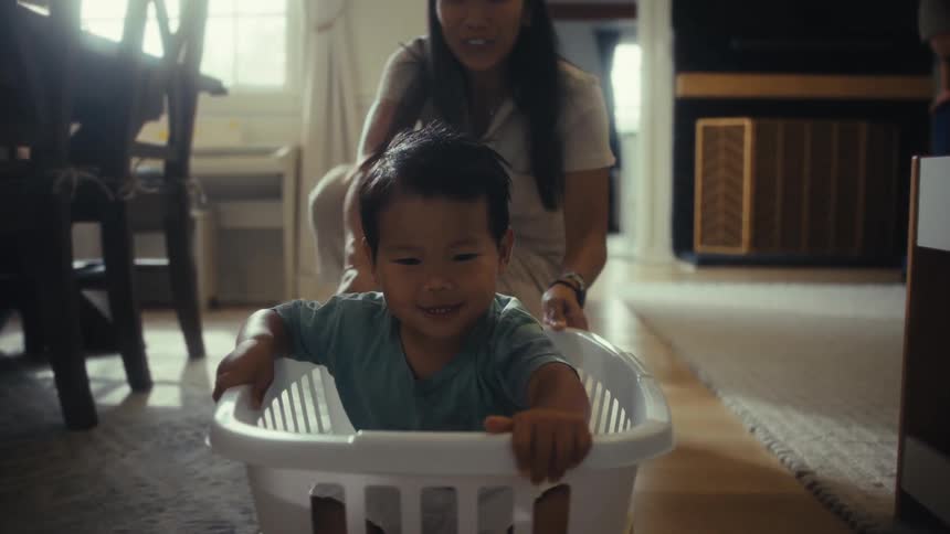 Young woman playfully drags a male toddler on a basket while both smile and an adult male holds another child on the background indoors during day time